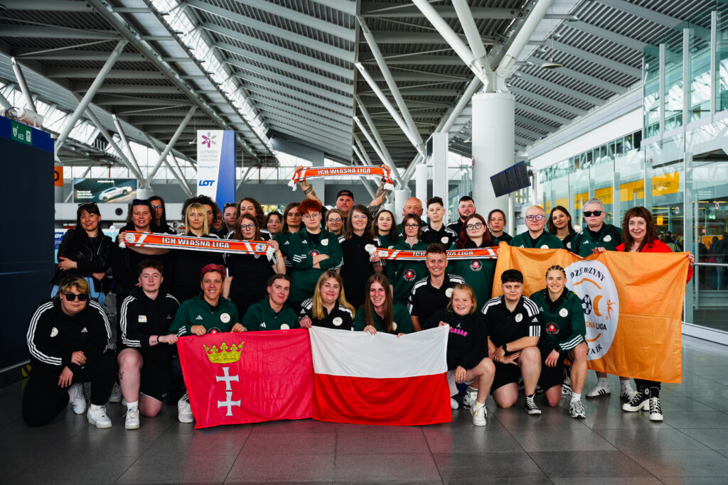 Grupa osób w sportowych strojach pozuje na lotnisku z flagami Polski, Gdańska i banerem ‘Ich Własna Liga’. Widoczne są szaliki z tym samym napisem. Tło stanowi przestrzeń terminalu